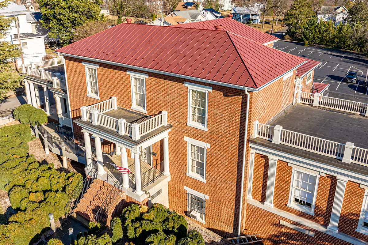 Large brick building with red metal roof and white trim, surrounded by greenery