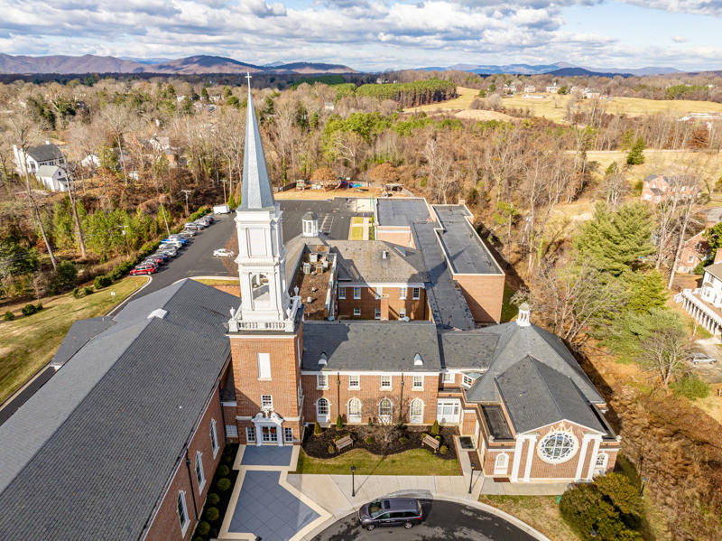 Aerial view of a brick church with a tall steeple surrounded by trees and hills