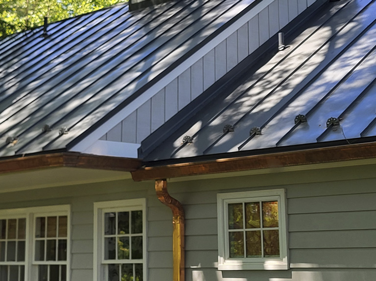 Gray house with a metal roof and leafless tree, situated next to a red brick building